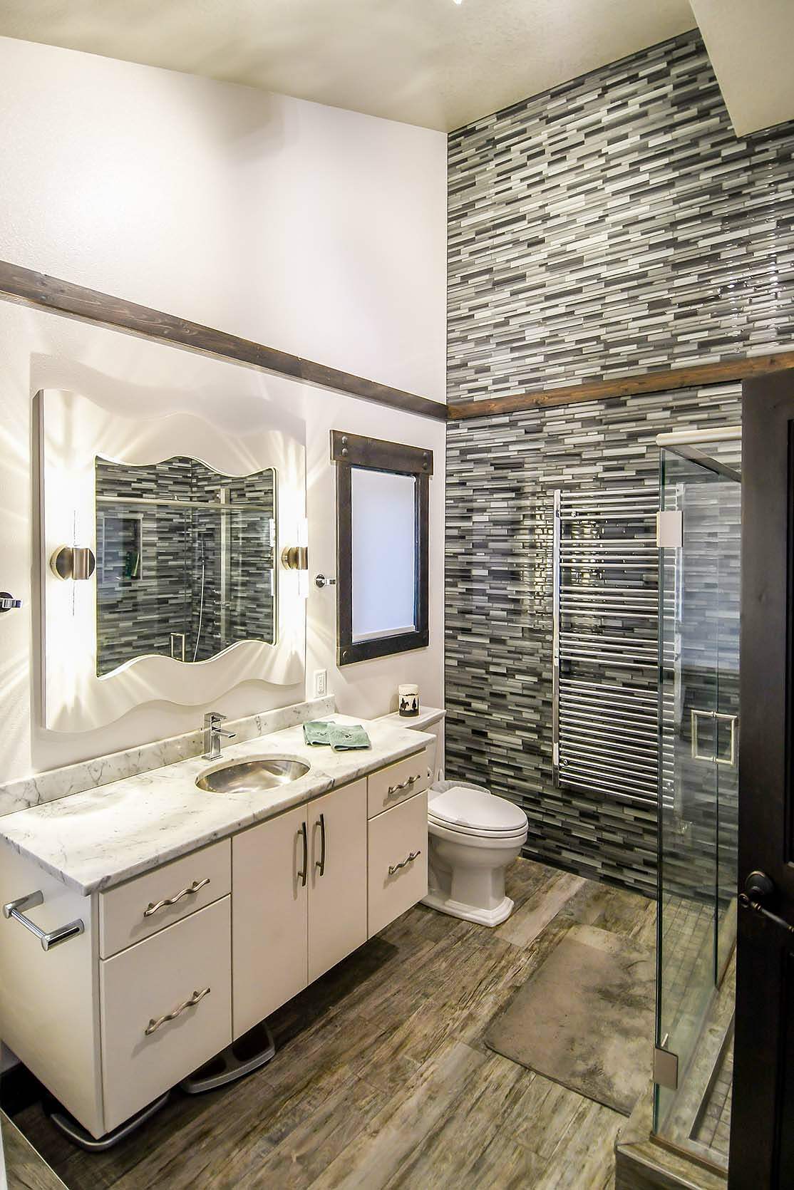 Modern bathroom interior featured in Lake & Home Magazine, March 2021, showcasing unique mirror shapes, a floating vanity with dual sinks, a textured gray accent wall, a glass-enclosed shower,
