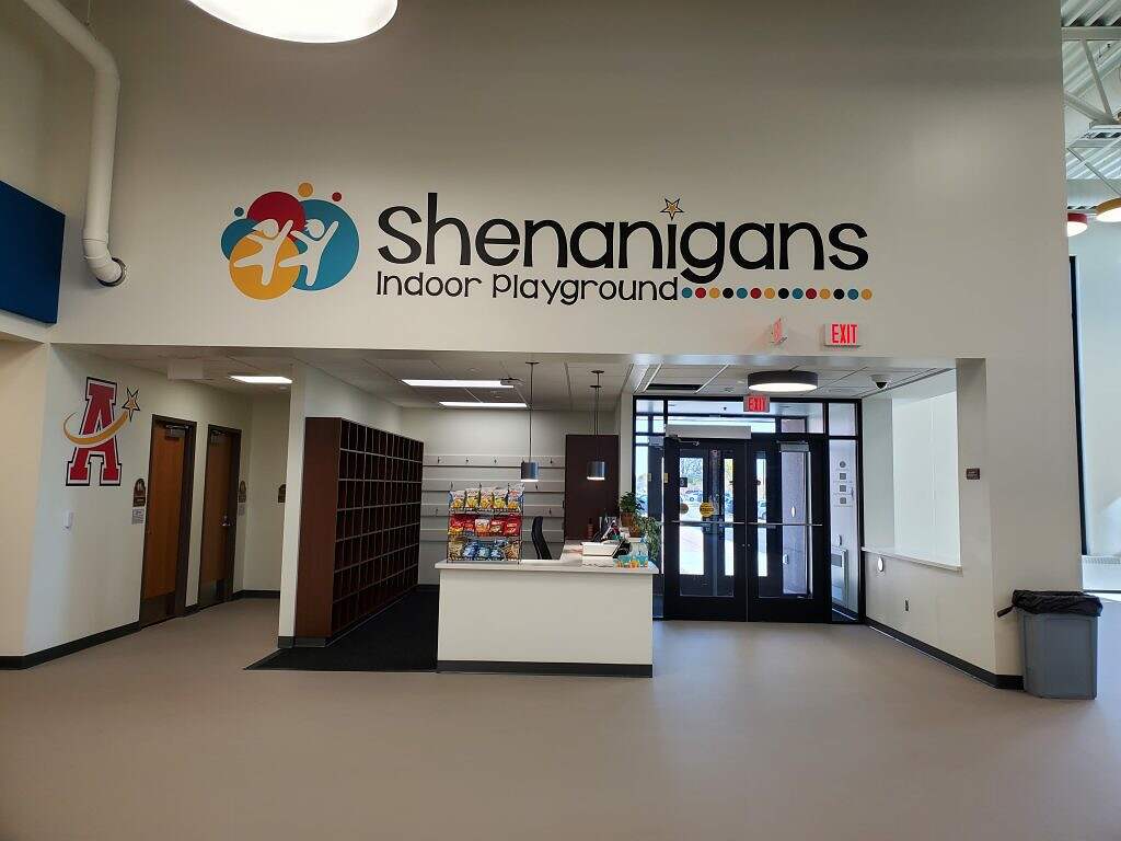 A brightly lit indoor playground with a sign that reads "Shenanigans," featuring playful and colorful graphics, inviting families to a fun and safe play area for children.