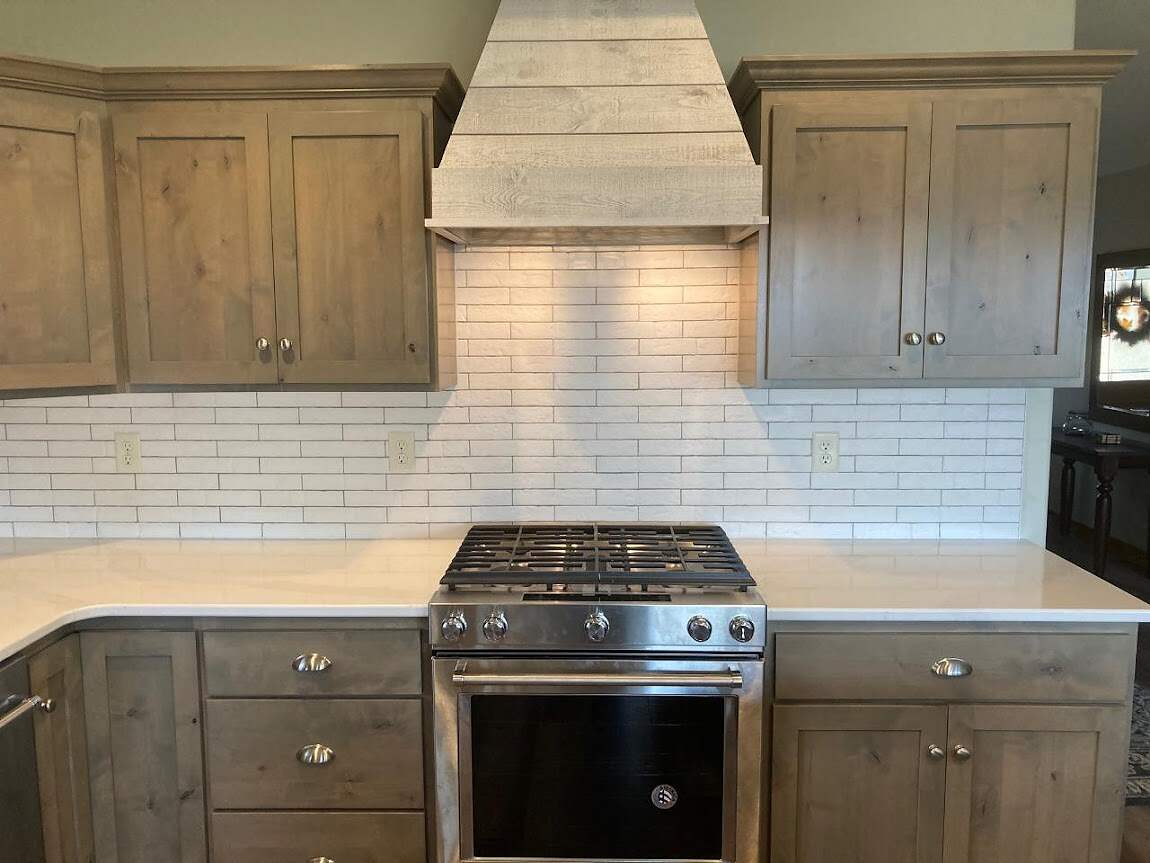 A modern kitchen with taupe-colored wooden cabinets, white subway tile kitchen backsplashes, and a stainless steel range with a matching vent hood.