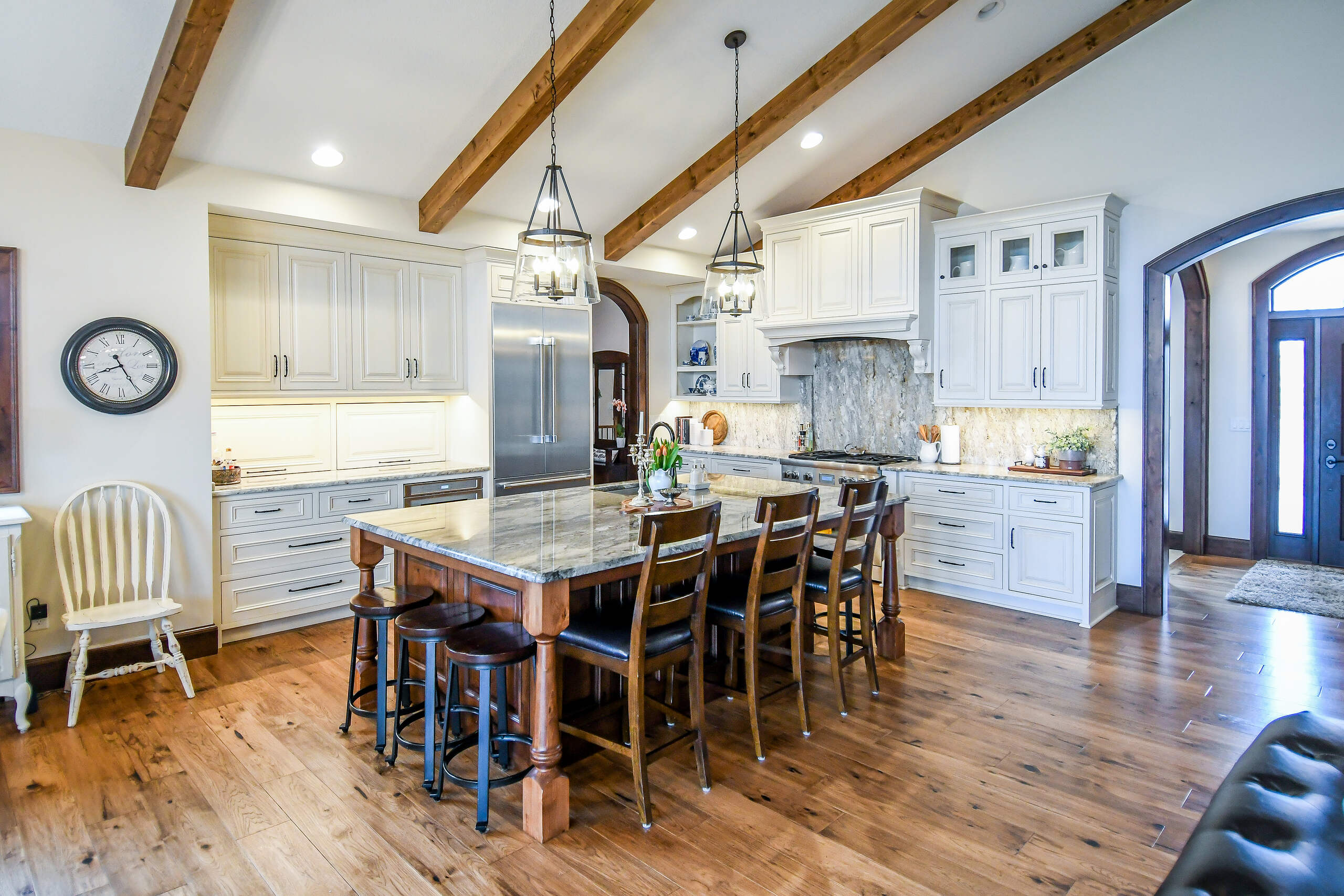 Spacious kitchen with rustic charm featuring white cabinetry, a stone backsplash, marble countertops, and an inviting wooden dining table set under elegant lighting.